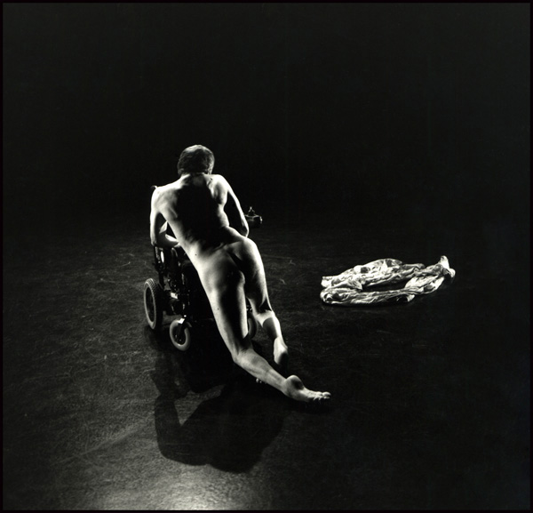 image of performer neil marcus nude climbing onto his wheelchair, in a dark theatre